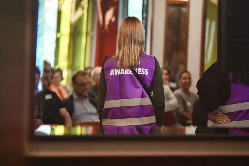 Eine Frau in Weste mit der Aufschrift "Awareness" steht vo einer sitzenden Menschengruppe.