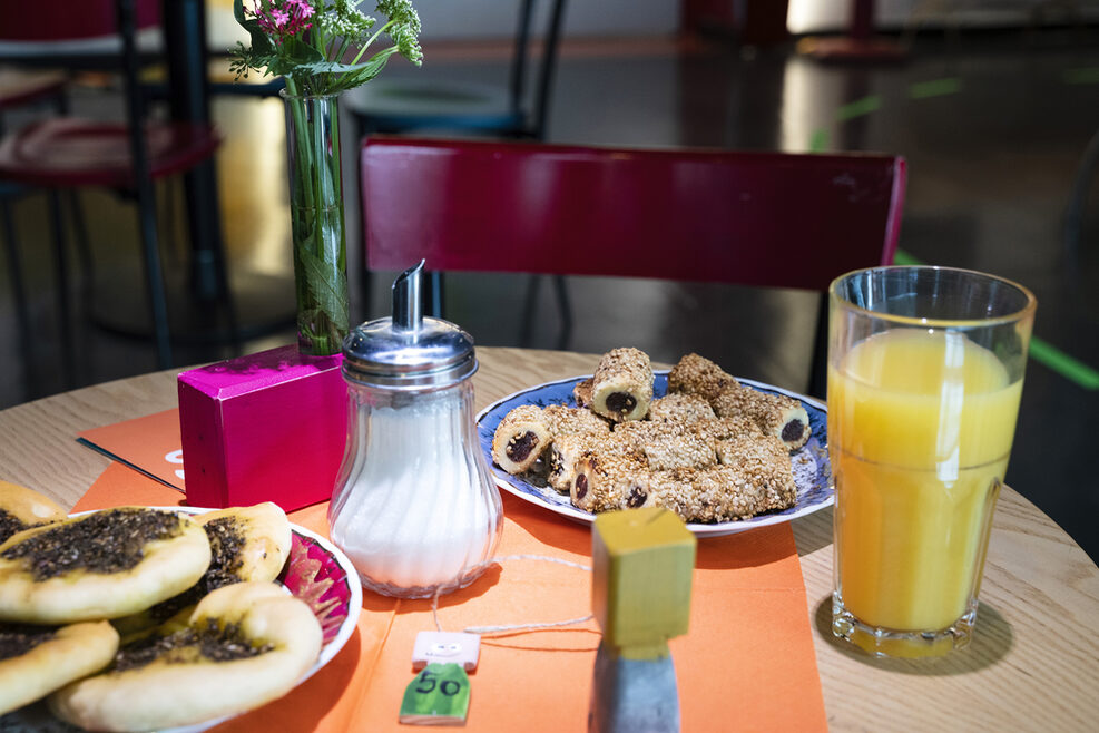 Auf dem Bild ist ein Tisch abgebildet. Auf dem Tisch steht ein Glas mit orangefarbenen Saft, einem Zuckerstreuer und zwei Tellern mit Gebäck darauf. Hinter dem Tisch steht ein roter Holzstuhl.