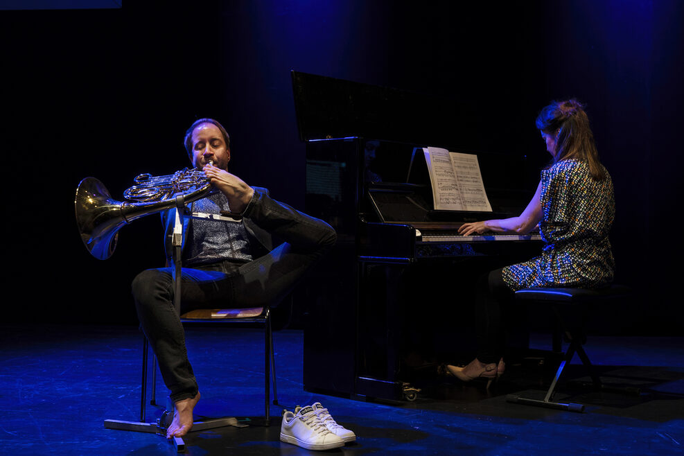 Auf dem Bild ist Felix Klieser zu sehen. Er sitzt auf einem Stuhl auf einer Bühne. Mit einem Fuß hält er sein Instrument Horn und spielt darauf. Rechts im Bild sitzt eine Frau an einem Klavier.