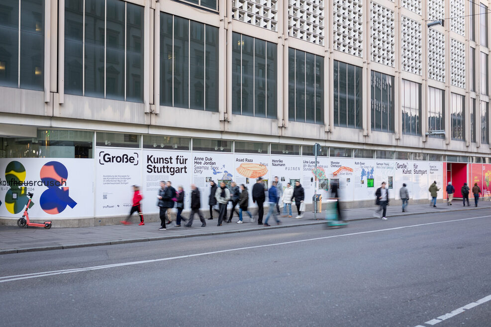 Die Fassade des ehemaligen Kaufhausgebäudes in Stuttgart, die mit Folien, auf denen Informationen zum Künstlerischen Großprojekt stehen, beklebt wurde. Vor dem Gebäude laufen mehrere Personen, die man nur verschwommen erkennen kann.