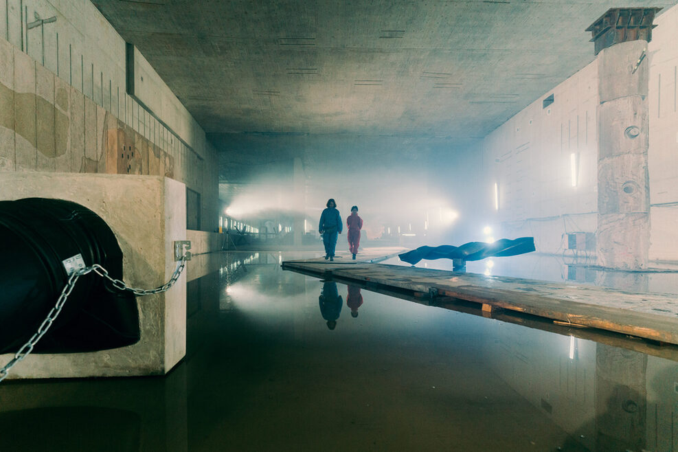 Auf der Baustelle Stuttgart 21 sind im diffusen Gegenlicht zwei Personen mit dem Rücken zum Betrachter auf einem Steg inmitten von Wasser zu sehen.