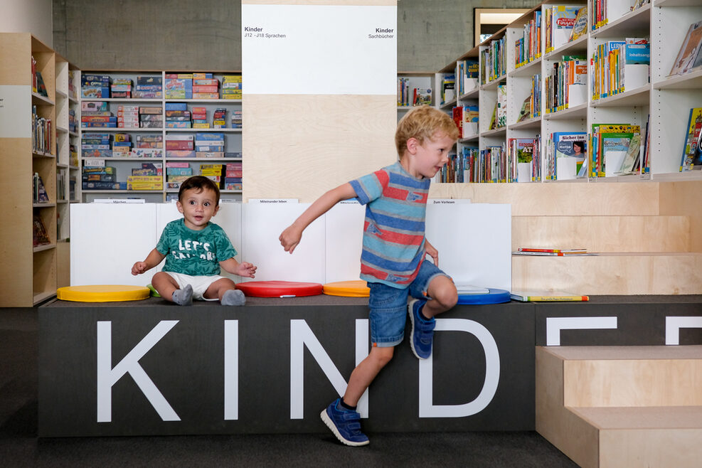 Zwei Kinder in der Stadtteilbibliothek Heslach: Ein Junge läuft durch die Regale, ein anderer sitzt auf dem Podest.