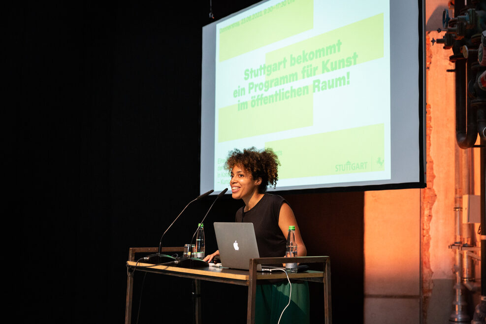 Tosin Stifel moderiert die Veranstaltung an. Im Hintergrund die Grafik "Stuttgart bekommt ein Programm für Kunst im öffentlichen Raum".