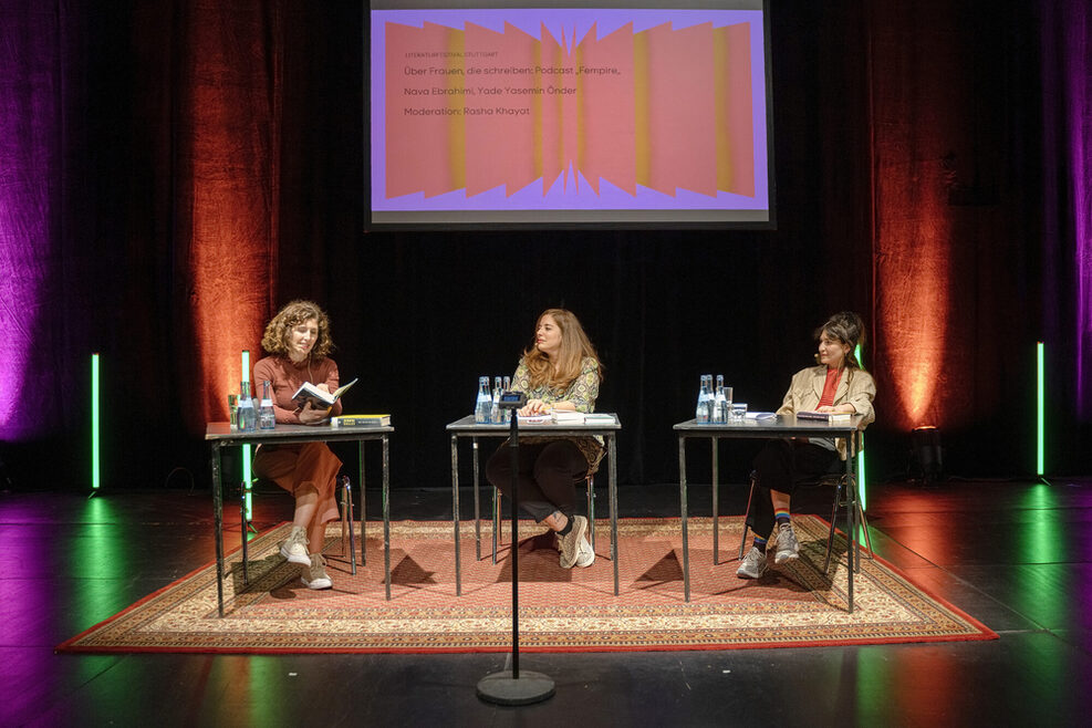 Drei Frauen sitzen an drei Tischen auf einer Bühne.