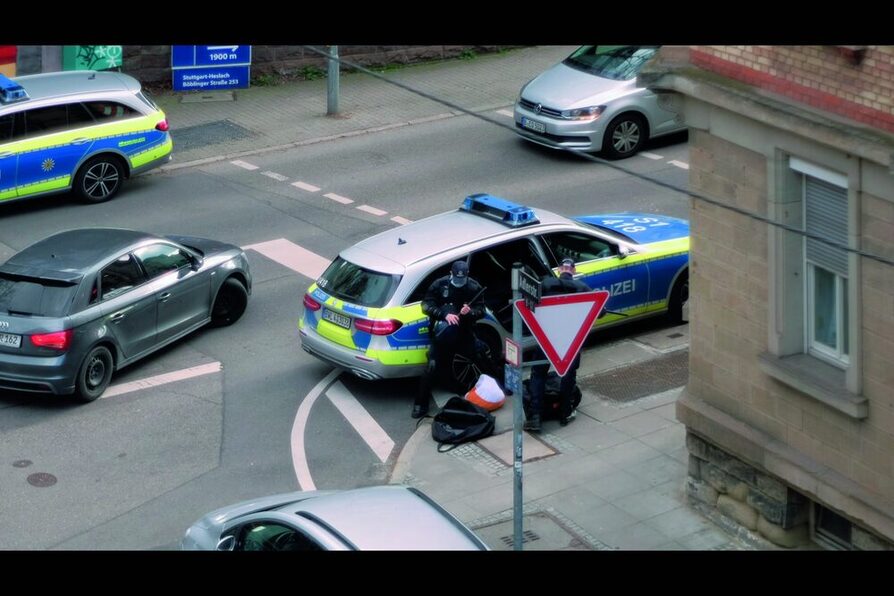 Eine Straßenkreuzung. Zwei Polizisten vor ihrem Auto (Videostill)
