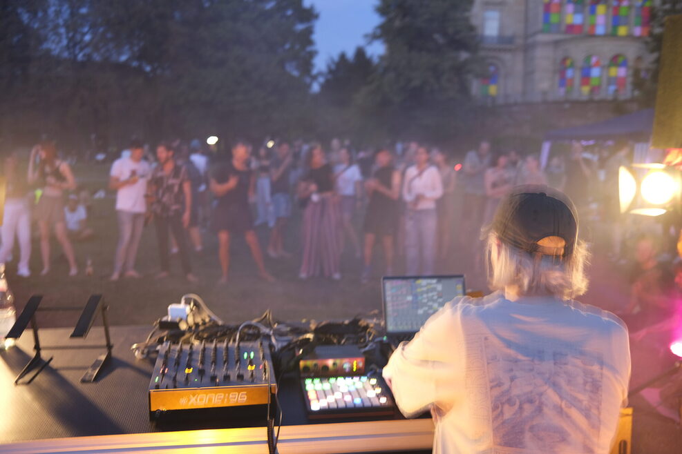 Blick auf ein Mischpult mit DJ Laima Adelaide von hinten. Im Hintergrund Personen im Park der Villa Berg.