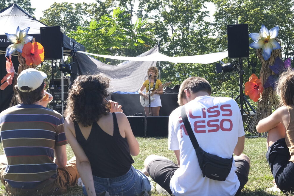 Im Hintergrund: Musikerin unter einem Zelt. Im Vordergrund sitzende Personen mit dem Rücken zum Betrachter.