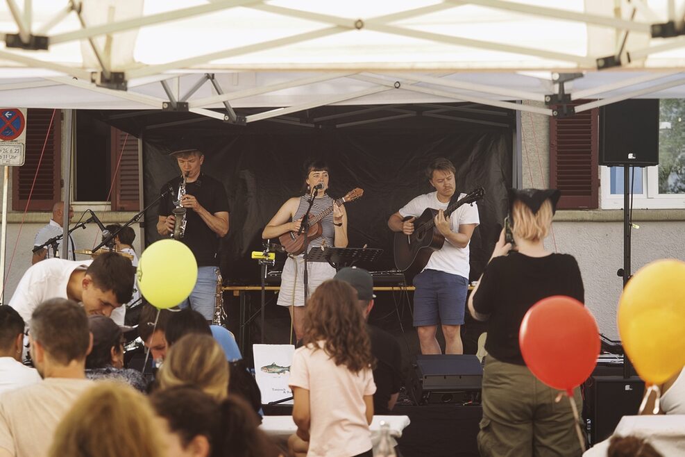 Drei Musiker der Band Tanjusch und Bande spielen unter einer überdachten Bühne vor Publikum. Zu sehen sind im Publikum Familien mit Kindern plus Luftballone.