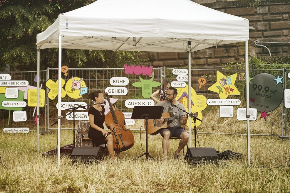 Eine Cellistin und ein Gitarrist sitzen unter einem Zeltdach vor einem Bauzaun,auf dem die Kunstausstellung GOOD TO KNOW X zu sehen ist,auf Stühlen im Gras und musizieren gemeinsam.