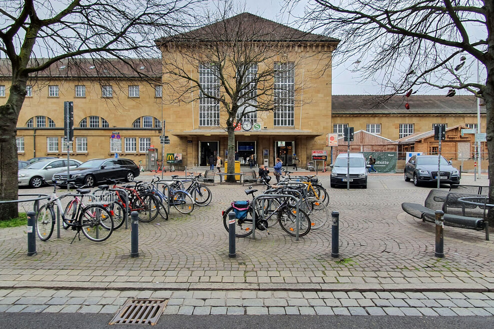Das Foto zeigt frontal den Vorplatz, auf dem viele Autos und Fahrräder parken. Im Hintergrund ist das Bahnhofsgebäude zu sehen.