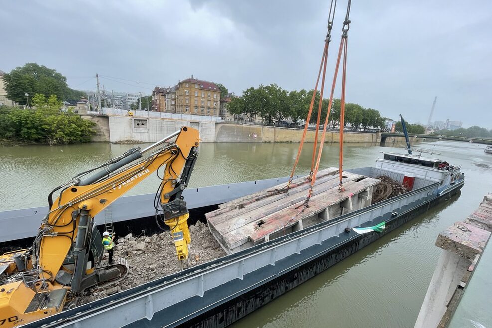 Ein großes Beton-Brückenteil wird auf ein Transportschiff verladen. Auf dem Schiff befindet sich außerdem ein gelber Kran, auf dessen Raupenkette ein Mann mit Bauhelm und Sicherheitsweste steht.
