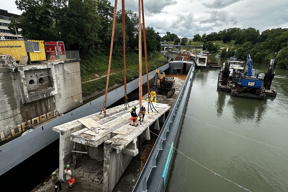 Das Teilstück des Brückenträgers wird auf das Schiff verladen. Einige Arbeiter befinden sich um das Stück herum und dirigieren den Kran.