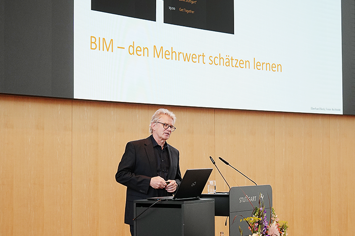 Herr Beck von Wabe-Plan Architektur hält am Pult des großen Sitzungssaals im Rathaus einen Vortrag zum Thema BIM. Auf der großen Leinwand im Hintergrund ist eine seiner Folien zu sehen. Der Titel lautet: BIM - den Mehrwert schätzen lernen.