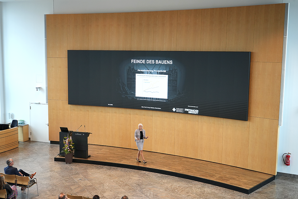 Frau Klemt-Albert hält im großen Sitzungssaal des Stuttgarter Rathauses einen Vortrag zum Thema BIM. Hinter ihr ist die große Leinwand mit einer Grafik zu sehen, vor ihr die Zuhörer in Stuhlreihen.