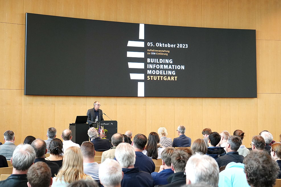 Bürgermeister Dirk Thürnau steht am Pult des großen Sitzungssaals des Stuttgarter Rathauses. Vor ihm sitzen in Stuhlreihen die Zuhörer, hinter ihm ist auf der Anzeigetafel vor schwarzem Hintergrund in Weiß und Gelb eine Grafik mit dem Titel Building Information Modeling Stuttgart und dem Datum 05. Oktober 2023 zu sehen.