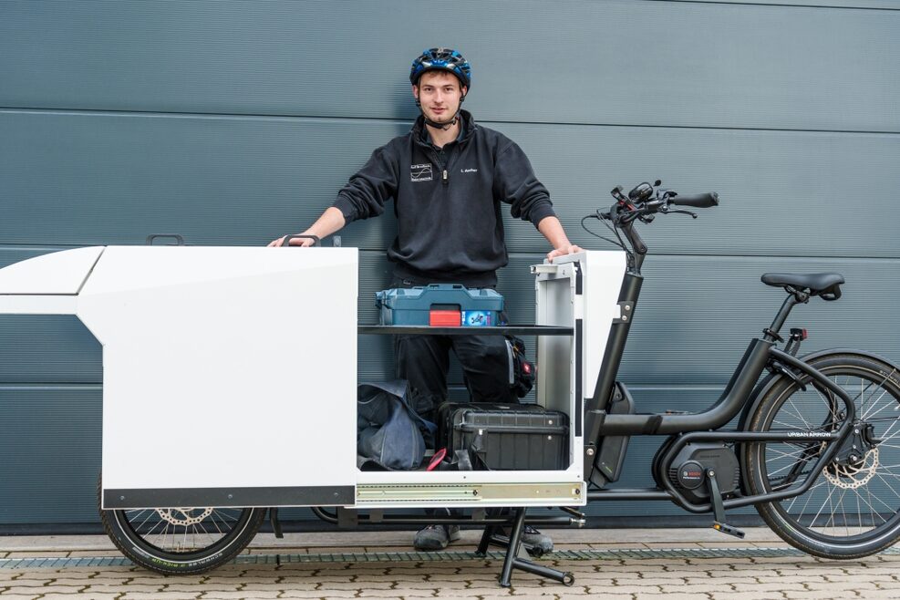 Ein junger Mann steht vor einer Hauswand und vor ihm ein Lastenrad.