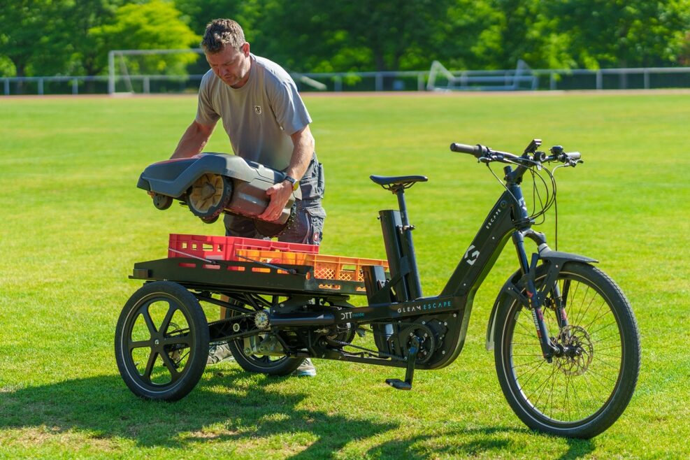 Ein Platzwart lädt ein auf einem Sportplatz stehendes Lastenrad mit einem Rasenmäher.
