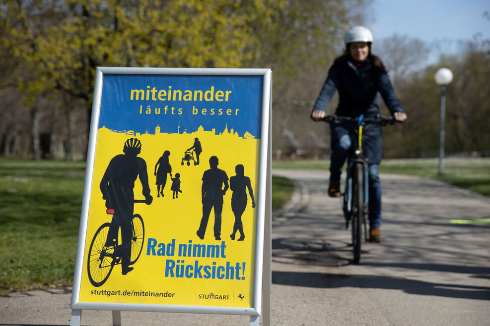 Das Plakat mit der Aufschrift "Rad nimmt Rücksicht" steht auf einem Fuß-und Radweg im Schlossgarten, eine Radfahrerin nähert sich im Hintergrund.