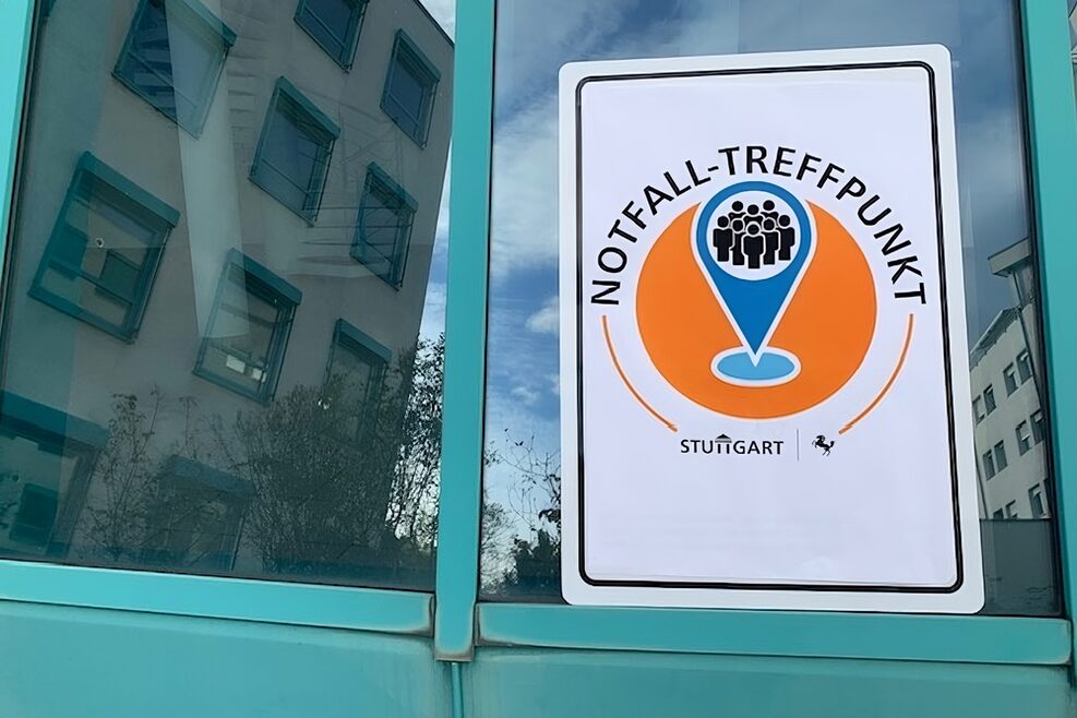 Schild mit der Aufschrift "Notfall-Treffpunkt" an einer Fensterscheibe, ein Kreis in Orange mit blauen Marker und Menschgruppe