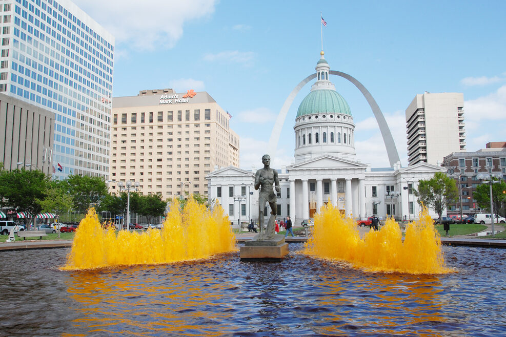 Brunnen mit einer Fontäne im Vordergrund, dahinter stehen ein historisches Gebäude, mehrere Hochhäuser und der Gateway Arch.