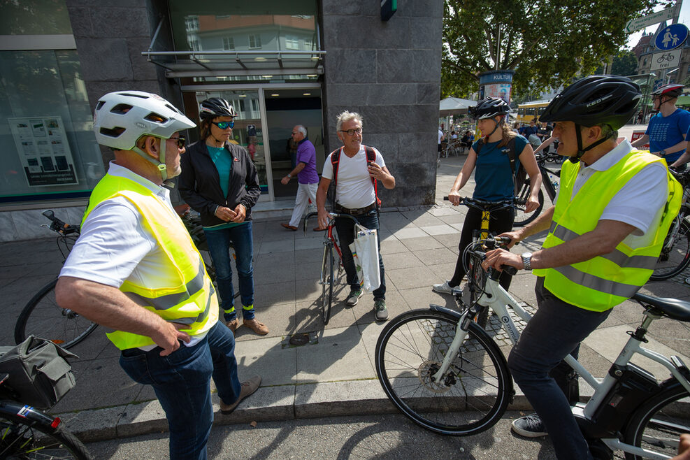 Eine Gruppe Menschen steht in einem Halbkreis. Links sind der Minister und rechts der Oberbürgermeister mit ihren Fahrrädern zu sehen. Im Hintergrund sieht man ihre Gesprächspartner, die ebenfalls vom Fahrrad gestiegen sind und sich mit ihnen unterhalten.