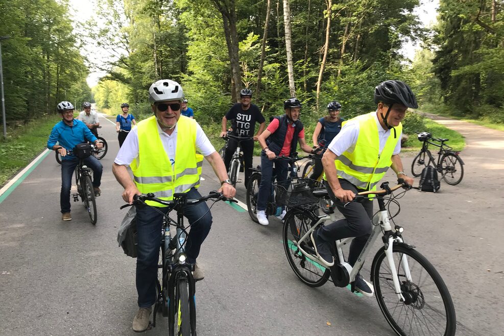 Eine Gruppe von Radfahrern ist auf einem asphaltierten Radweg durch ein Waldgebiet unterwegs. Im Vordergund sind Baden-Württembergs Verkehrsminister Winfried Hermann (vorne links) und Stuttgarts Oberbürgermeister Dr. Frank Nopper (vorne rechts) zu sehen.