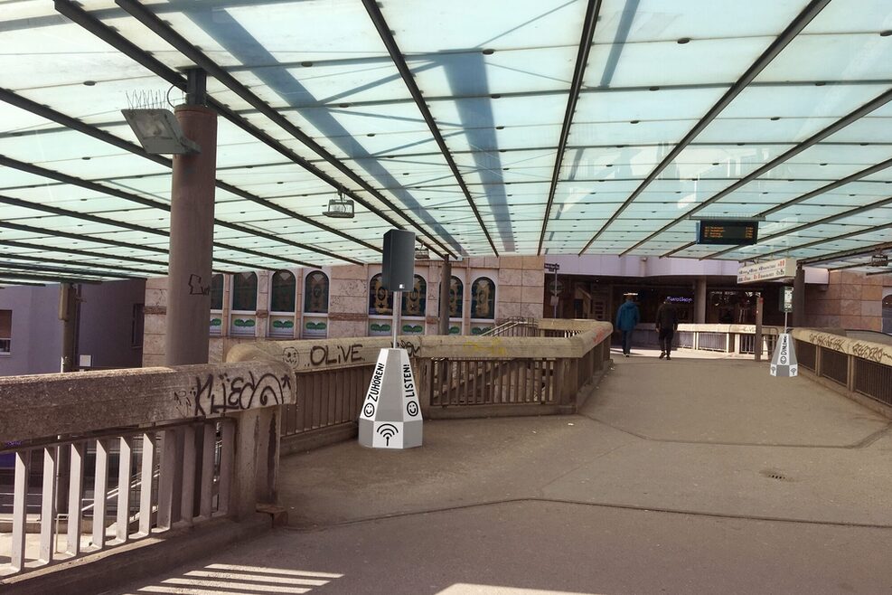 Die Ansicht des Martin-Mayer-Stegs oben mit dem Dach des Bahnsteigs darüber. Ein Lautsprecher steht auf dem Steg.