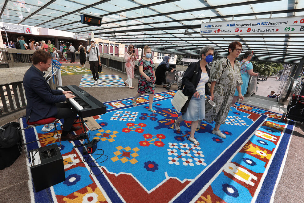 Installation "Den Teppich ausrollen" am Martin-Mayer-Steg. Zu sehen sind bunte Teppiche und Menschen, dir darüber laufen Richtung Gleise.