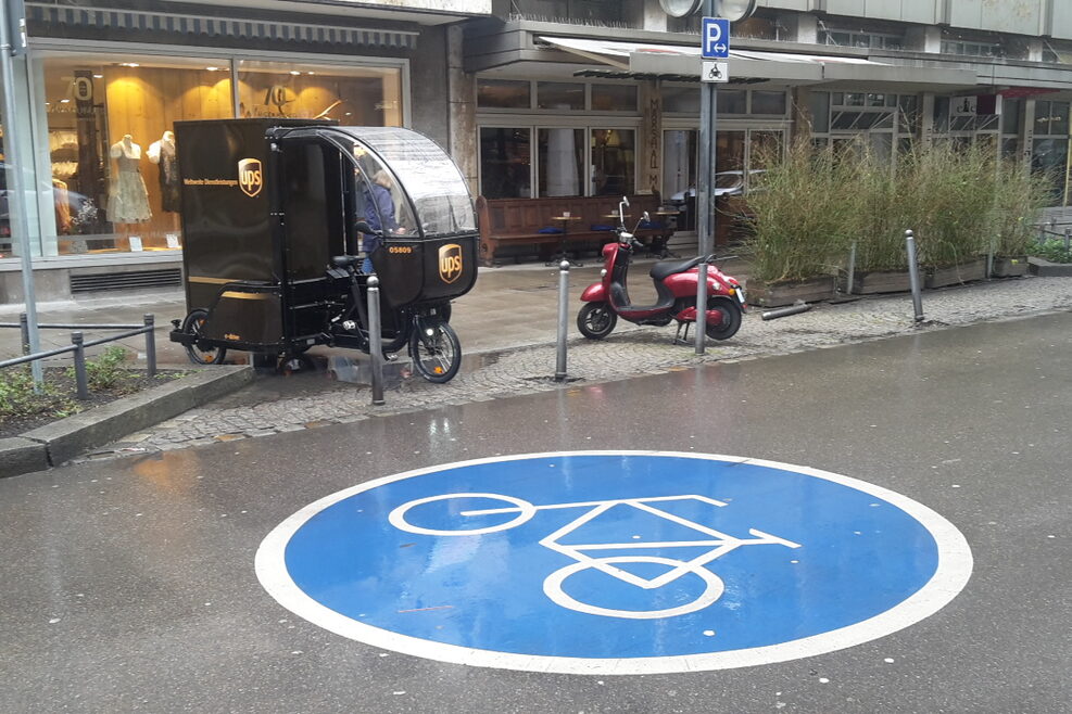 In der Eberhardstraße ist auf dem Asphalt ein großes Zeichen für Fahrradweg aufgebracht. Im Hintergrund steht am Rande des Fußwegs auf einer kleinen Parkzone ein elektrischen Dreirad eines Paketdienstes. Es regnet.