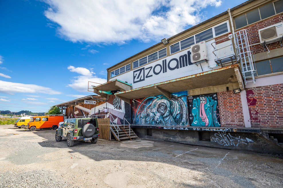 Buntes Backsteingebäude mit der Aufschrift „Zollamt“. Davor parken Autos.