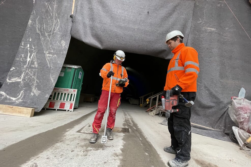 Zwei Arbeiter vermessen Fahrbahn, im Hintergrund ist ein Tunneleingang zusehen.