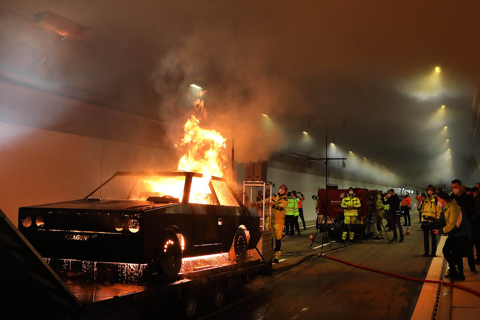 Eine Autoatrappe steht in einer Tunnelröhre. Aus dieser schlagen Flammen. Im Hintergrund sind Feuerwehrmänner und Journalisten zu sehen. An der Decke des Tunnels sieht man die Rauchentwicklung.