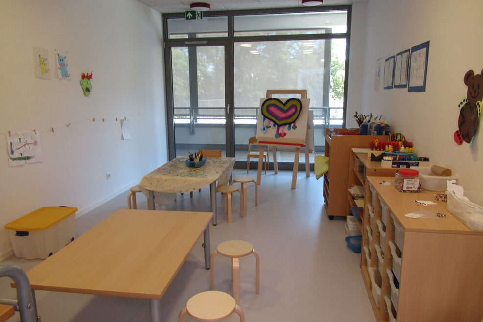 Atelier der Kita Rostocker Straße in Bad Cannstatt mit einer Staffelei hinten im Raum, Tischen in der Mitte und Regalen, in denen verschiedene Stiftarten und Farbmaterialien untergebracht sind.