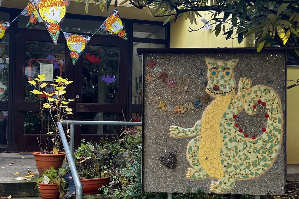 Eingangsbereich der Kita Bottroper Straße in Bad Cannstatt mit einem selbstgestalteten Mosaik, das einen Dinosaurierer abbildet. In das Gebäude führen ein paar Treppenstufen.