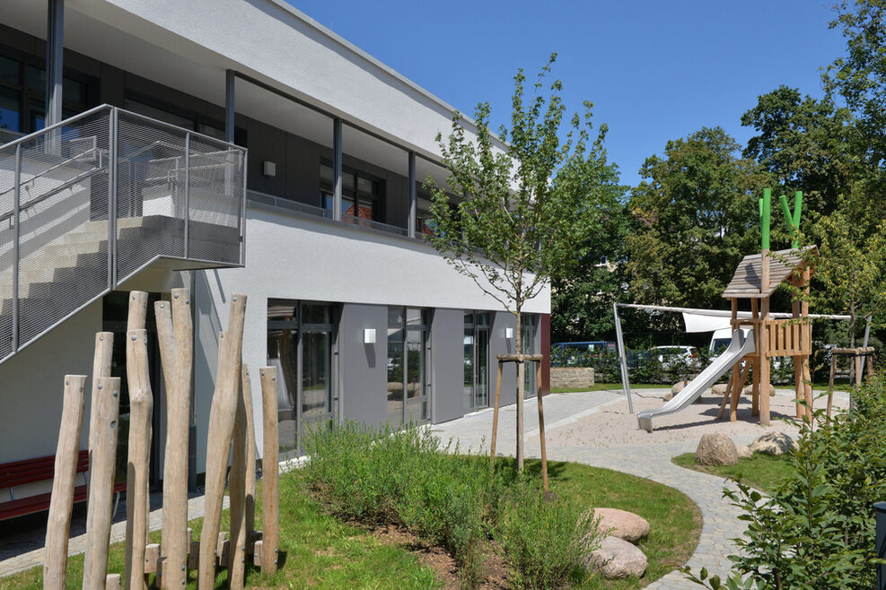 Der weitläufige Garten der Kita Rostocker Straße in Bad Cannstatt mit Grünflächen, einem Sandbereich mit Klettergerüst und Sitzmöglichkeiten.