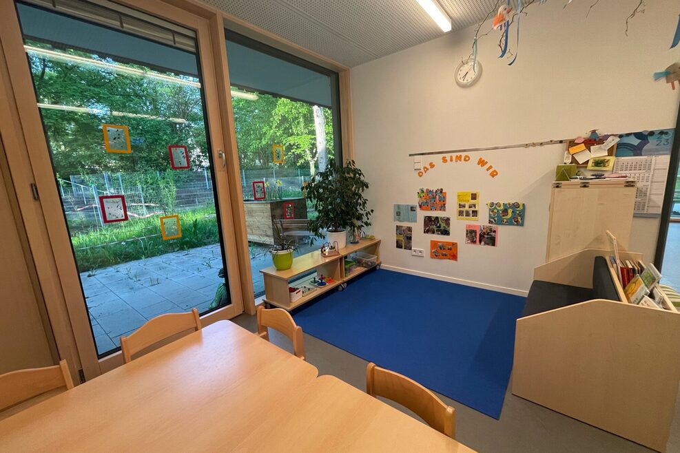 Kleinkindgruppenraum mit Tisch im Vordergrund und Sitzplatz mit Büchern.
