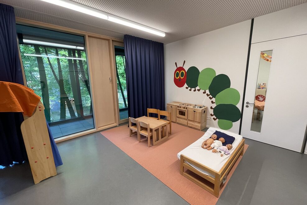 Kita Raum mit Tisch und Stühlen, Küche und Bett. An der Wand ein Bild mit einer Raupe.