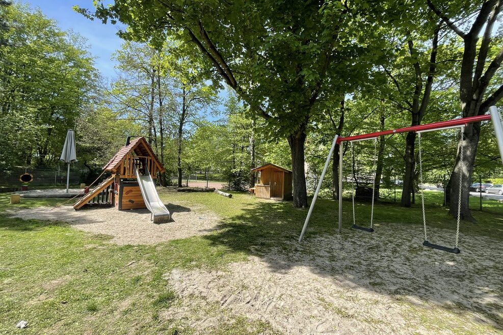 Spielplatz mit Schaukel und Rutsche unter Bäumen.