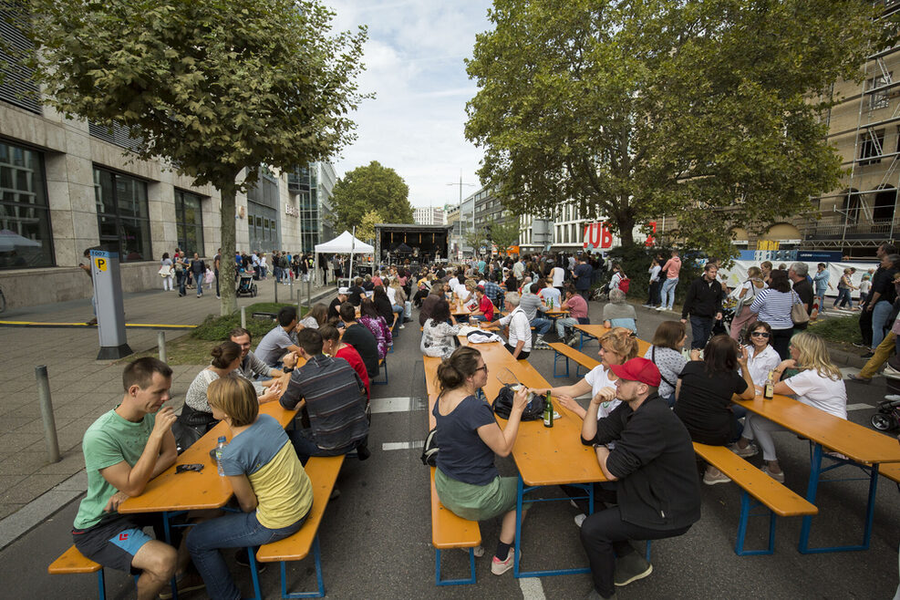 Menschen sitzen an Bierbänken auf der Theodor-Heuss-Straße beim Essen und Trinken.