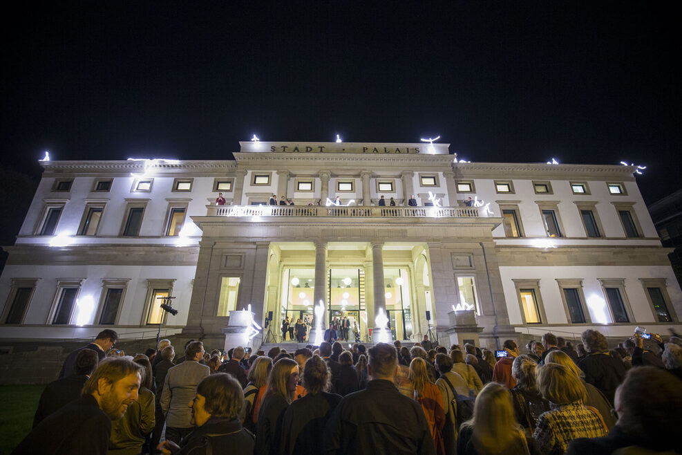 Mehrere Menschen stehen bei Nacht vor dem Stadtpalais, das hell beleuchtet ist.