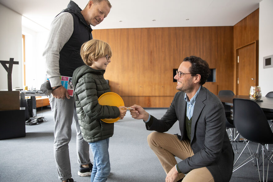 Stuttgarts Erster Bürgermeister kniet in seinem Büro vor einem kleinen Jungen und seinem Vater und unterhält sich.