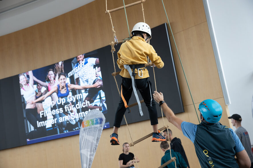 Ein Kind klettert mit einem Seil gesichert eine Strickleiter hoch, die von einem Erwachsenen festgehalten wird. Im Hintergrund hängt die digitale Leinwand des Rathauses an der Wand.