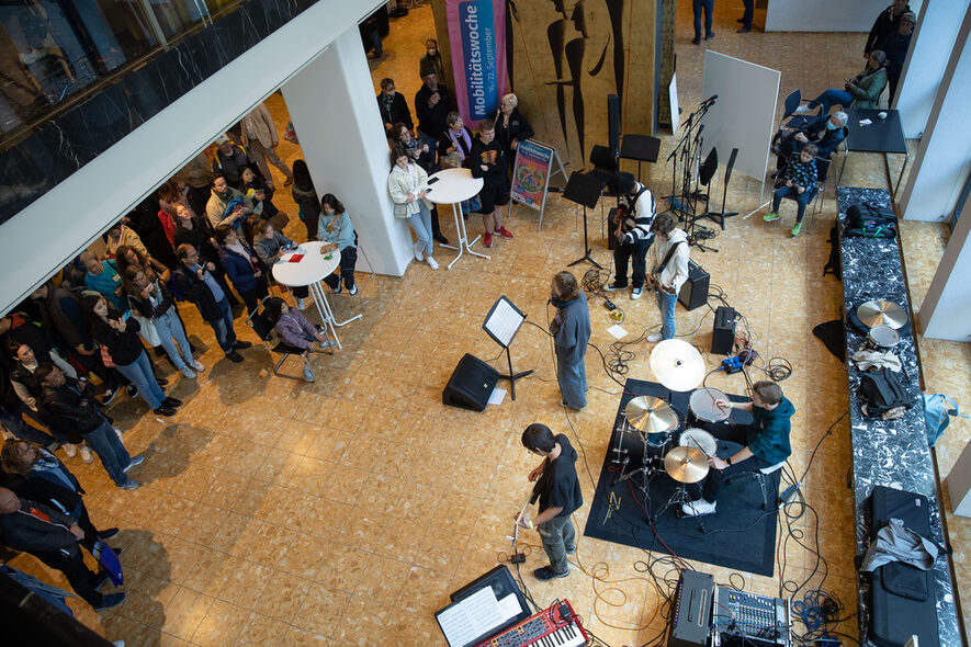 Blick von oben auf eine Band im Foyer. Links im Bild stehen Zuschauer und hören der Musik zu.