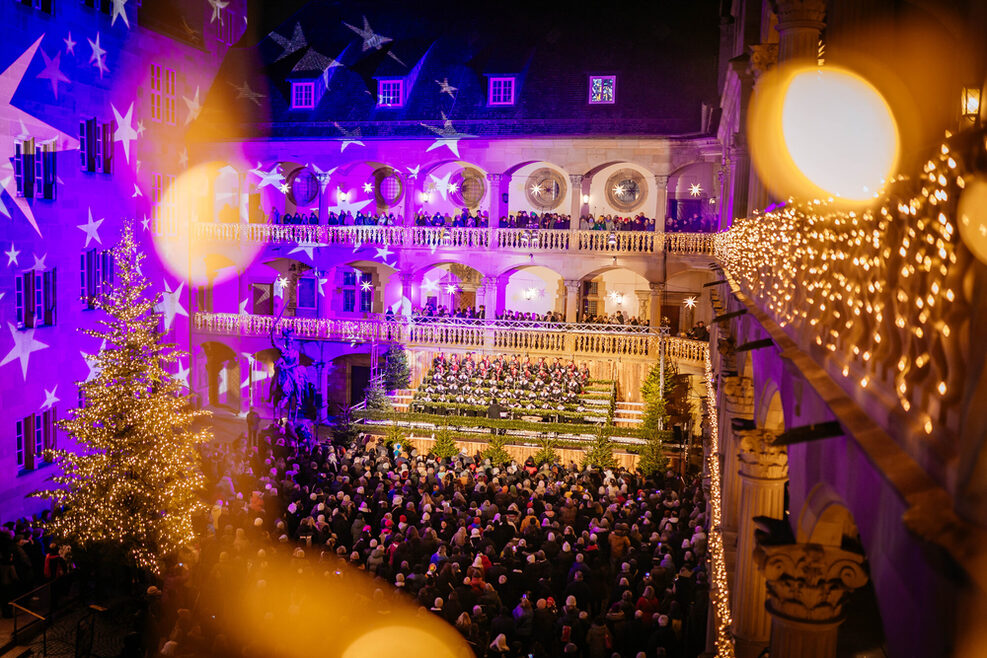View of the festively decorated courtyard of the Old Palace in Stuttgart at the opening ceremony of the Christmas market with a choir, many spectators and projected stars on the walls.
