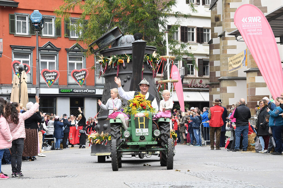 Festwagen wird von einem Traktor gezogen, der Fahrer sowie zwei kleine Mädchen winken vom Traktor.