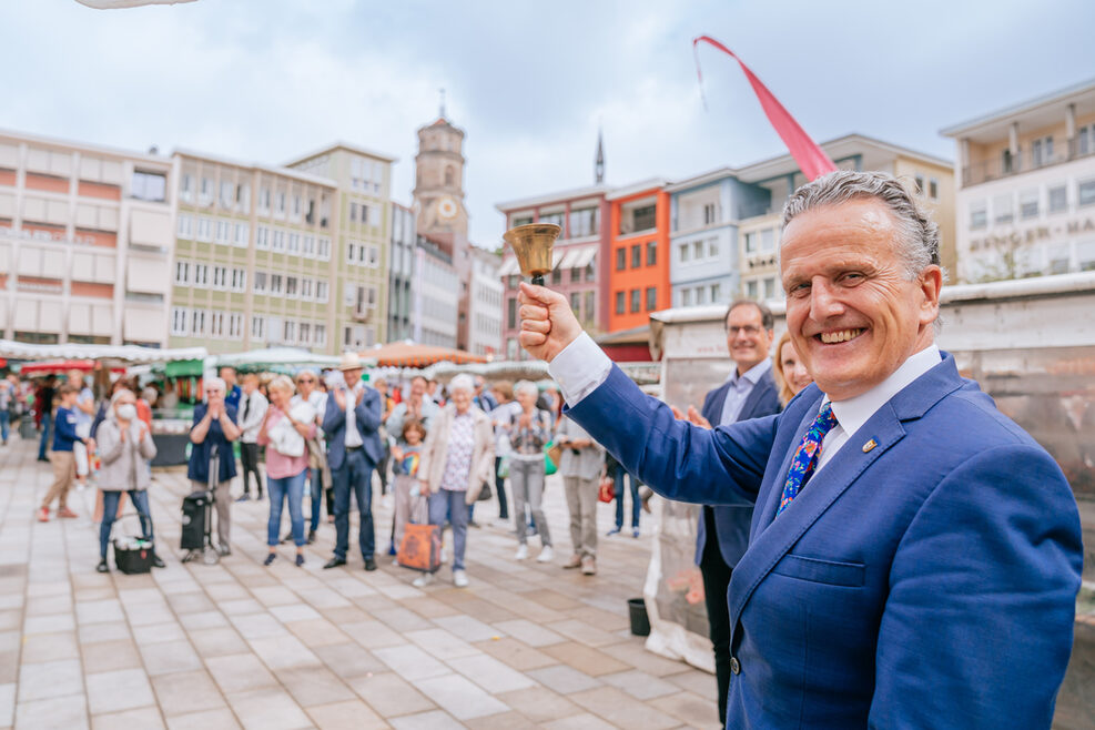 Der Oberbürgermeister steht auf dem Marktplatz und läutet eine Glocke. Im Hintergrund applaudieren menschen und es sind Marktstände zu sehen.