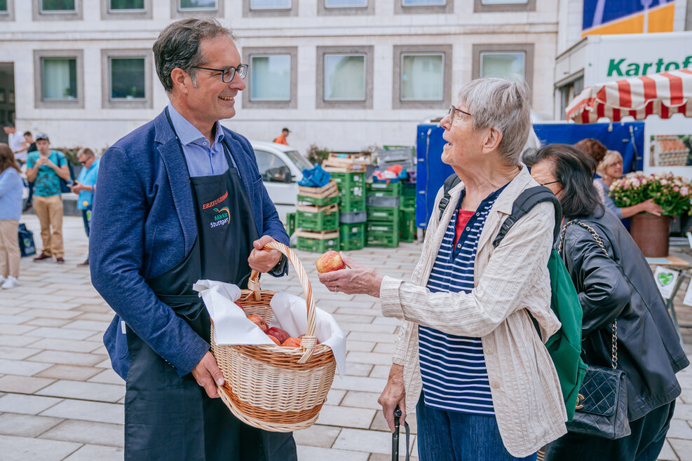Bürgermeister Clemens Maier hat einer älteren Dame einen Apfel überreicht. In den Händen hält er einen Korb mit weiteren Äpfeln. Im Hintergrund sind Teile des Stuttgarter Rathauses zu erkennen.