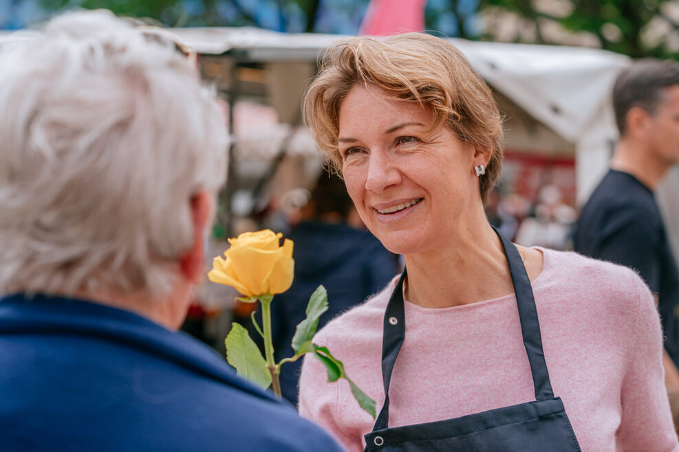Alexandra Sußmann überreicht lächelnd einer Dame eine gelbe Rose. Im Hintergrund sind Marktstände zu sehen.
