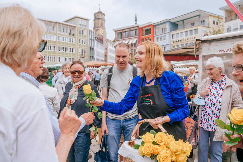 Bürgermeisterin Isabel Fezer überreicht einem älteren Herren eine gelbe Rose. Es stehen Menschen um sie herum. Im Hintergrund sind Marktstände und Häuser zu sehen.