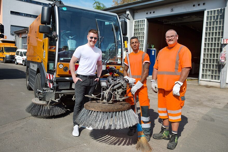 Drei Personen stehen vor einer orangefarbenen Kehrmaschine.